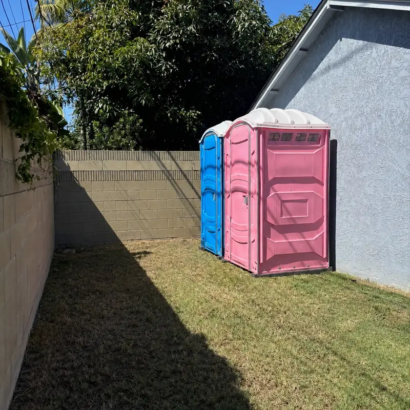 Pink and blue event porta potties set on grass along a backyard fence